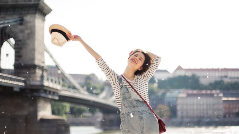 Content female with closed eyes standing on waterfront near bridge with straw hat and raised arm with hand behind head in city on blurred background