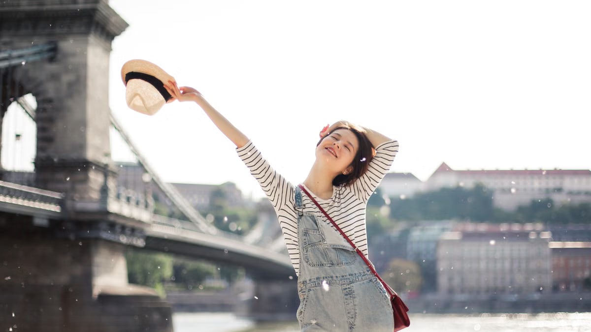 Content female with closed eyes standing on waterfront near bridge with straw hat and raised arm with hand behind head in city on blurred background