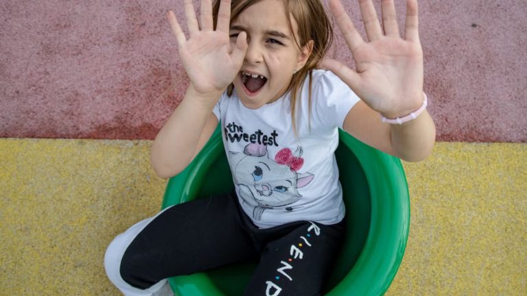 A joyful girl with brown hair sitting in a green chair raises her hands playfully.